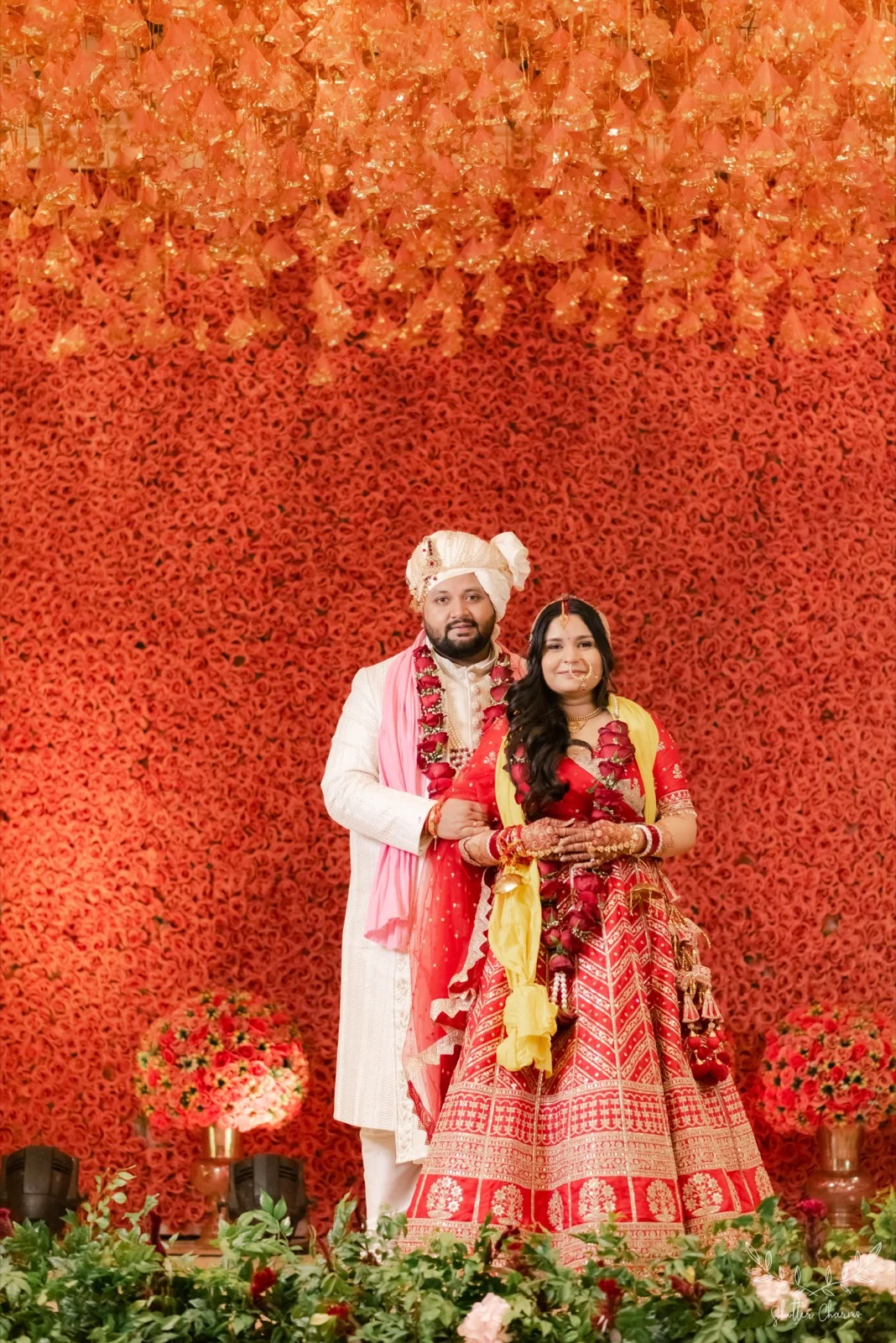 Newlyweds with rose garlands posing against red floral backdrop