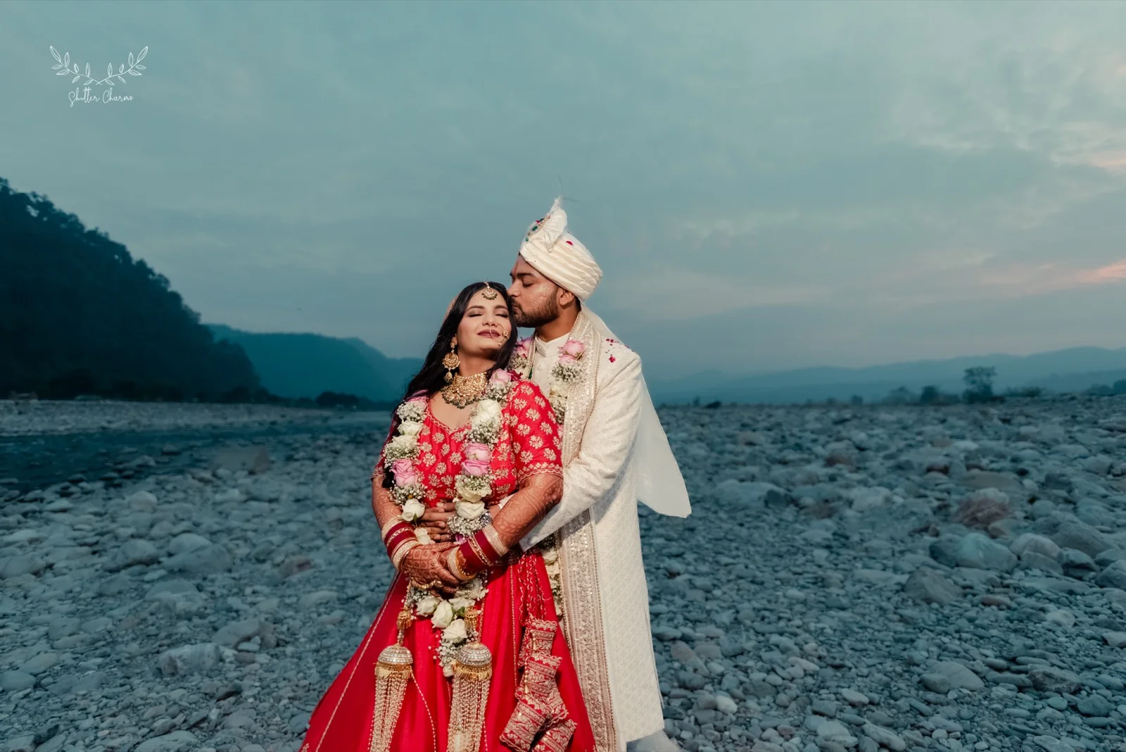 Groom kissing bride's forehead on a scenic riverbed at dusk