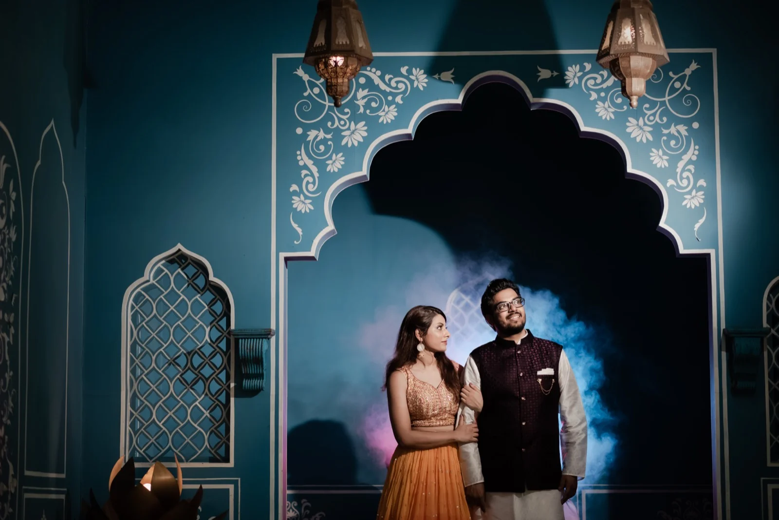 Couple in ethnic wear posing by blue jharokha backdrop with lanterns